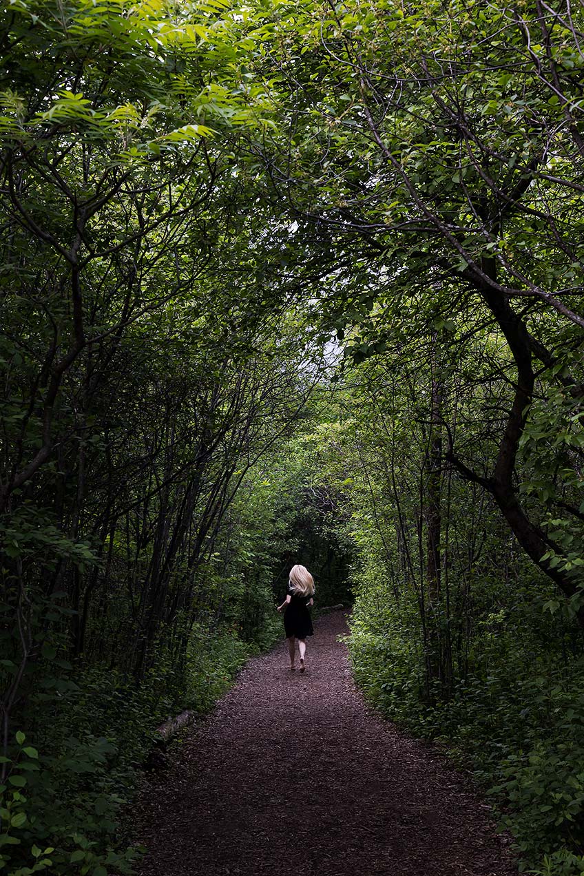 Le Repos de la Guerrière - Promenons-nous dans les bois - Woman in a black dress running down a forest path covered by arching trees.
