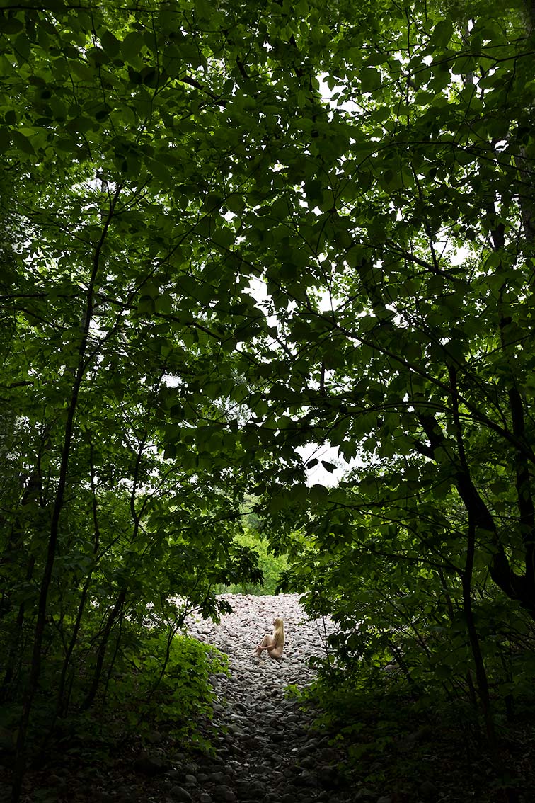 Le Repos de la Guerrière - Embryo - Woman sitting on a rocky slope as seen through the forest canopy.